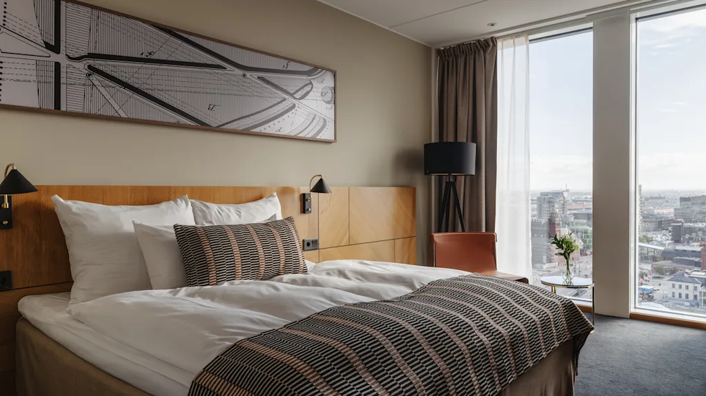 Et moderne hotellrom med en komfortabel seng, et stort vindu som tilbyr byutsikt, og et nattbord med friske blomster.