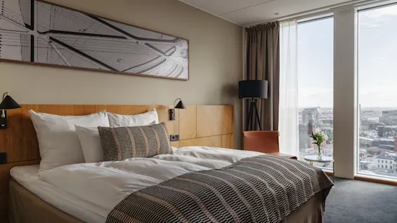 Et moderne hotellrom med en komfortabel seng, et stort vindu som tilbyr byutsikt, og et nattbord med friske blomster.