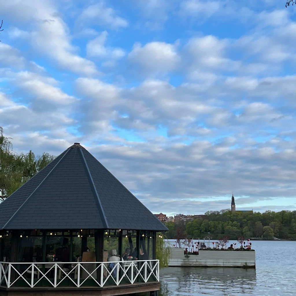 Människor som äter i en mysig paviljong vid vattnet under en molnig blå himmel.