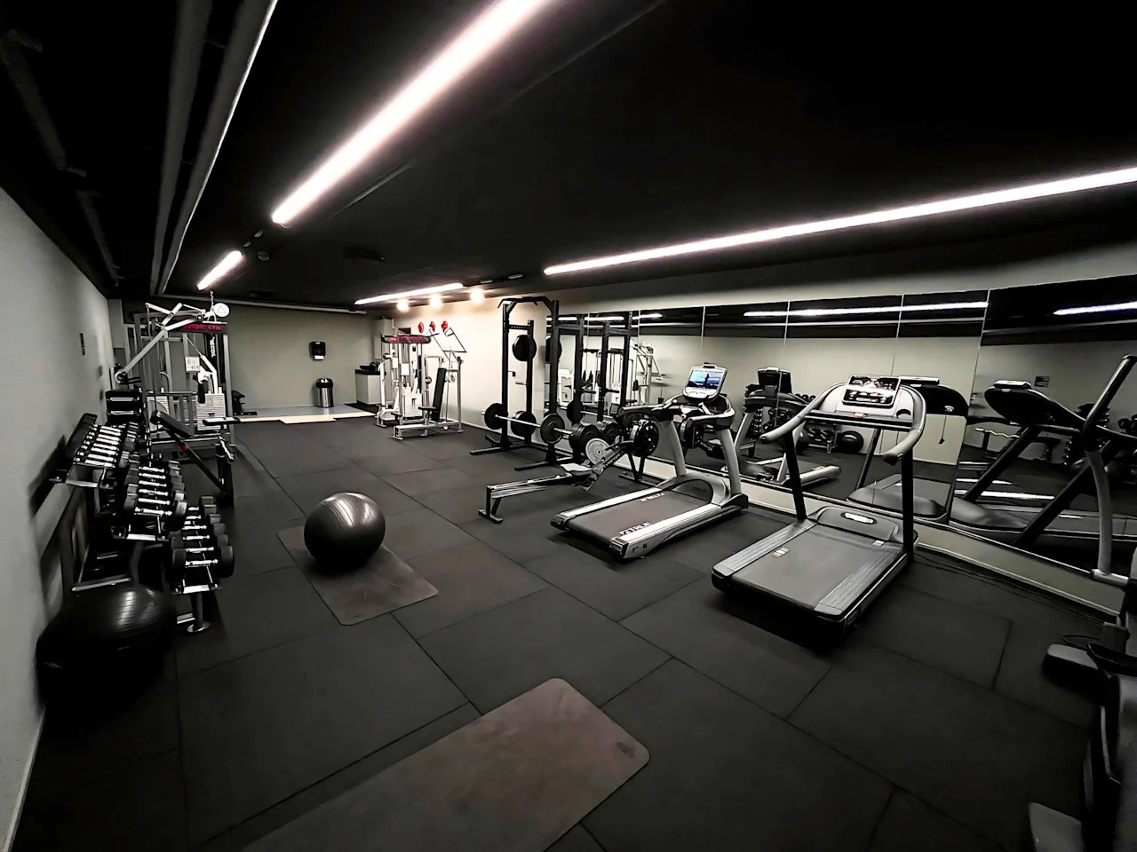 Treadmills line the mirrored wall, stationary in an empty gym. Dumbbells and exercise balls rest on padded floors under bright ceiling lights.