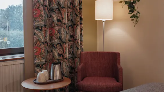A cozy corner at Home Hotel Kung Oscar with a patterned curtain, a red armchair, and a small table with a kettle.
