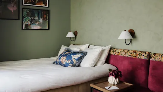 A bed with white linens and a floral pillow rests against a green wall. Above are framed artworks. A small table with a vase and flowers is nearby.