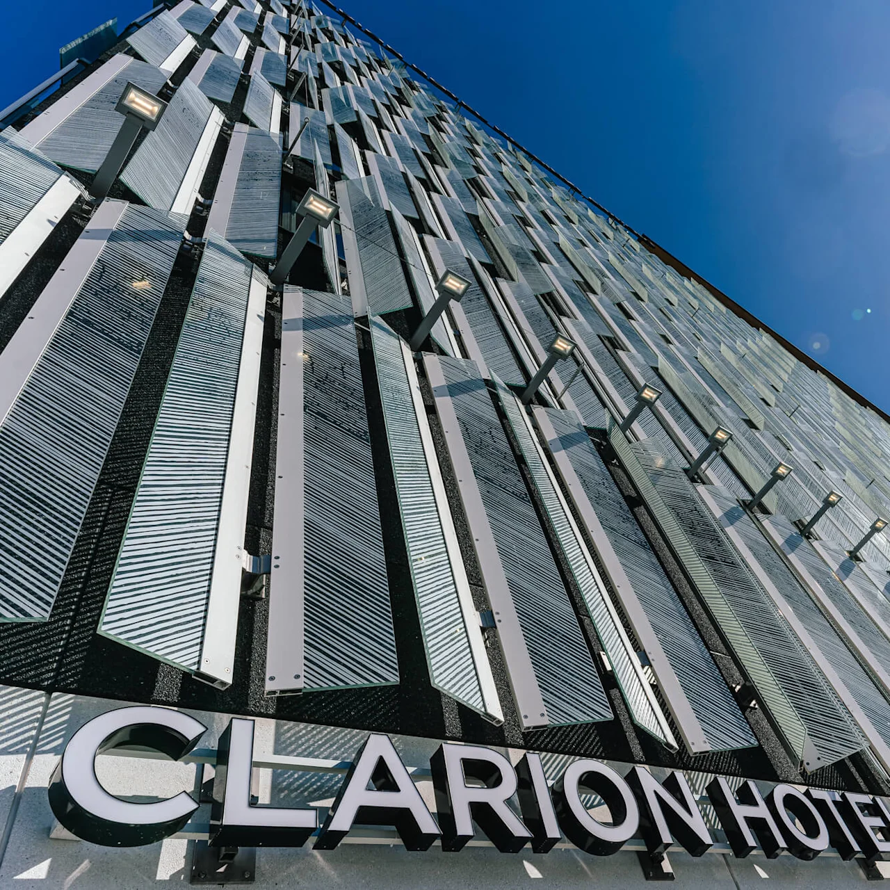 A modern building exterior with vertical metal panels reflecting sunlight under a bright blue sky. Text reads "CLARION HOTEL HELSINKI AIRPORT".