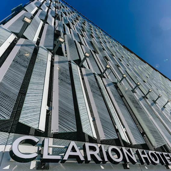 Modernin rakennuksen ulkopinta, jossa on pystysuuntaiset metallipaneelit heijastamassa auringonvaloa kirkkaan sinisen taivaan alla. Tekstissä lukee "CLARION HOTEL HELSINKI AIRPORT".