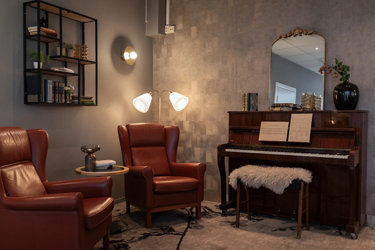 An inviting lounge at Home Hotel Grand Bodø featuring comfortable leather armchairs, a piano with sheet music, and warm lighting.