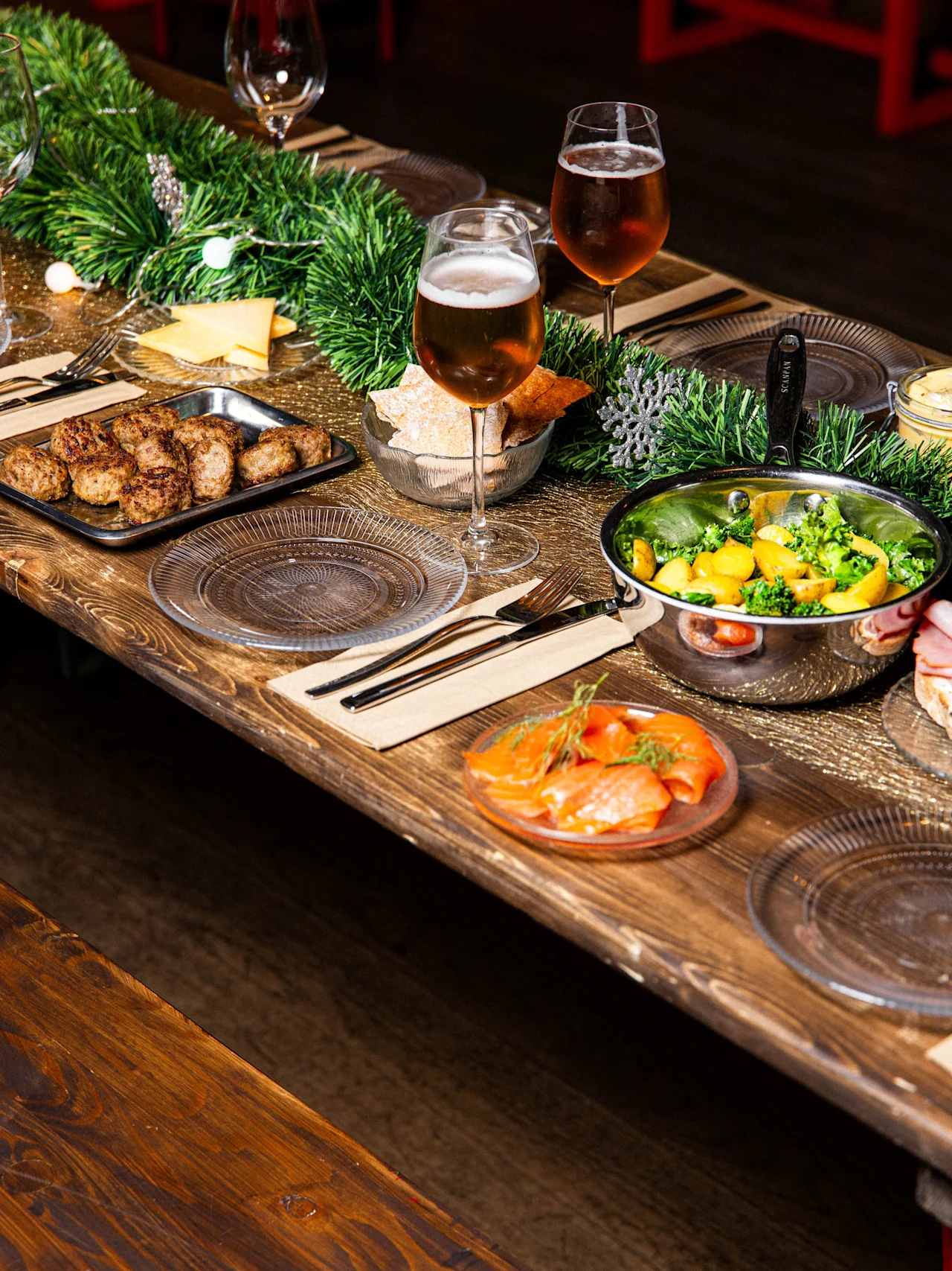 A wooden table displays prepared food, such as vegetables, meat, and smoked salmon, alongside beer glasses. A green garland decorates the setting, creating a festive dining atmosphere.