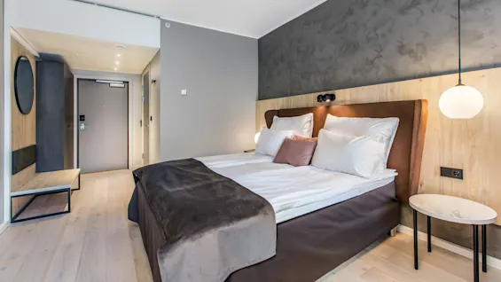 A neatly made double bed with white and brown bedding sits against a textured headboard. To the right, a round hanging light and a small side table enhance the modern room.