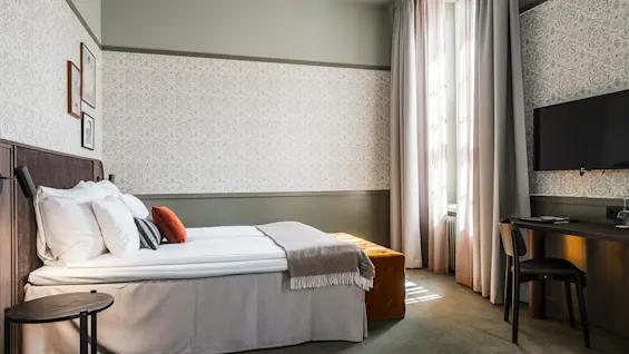 Ett snyggt bäddad säng i ett välbelyst hotellrum med dekorativa kuddar, intill ett arbetsutrymme med en svart stol och en TV på väggen.