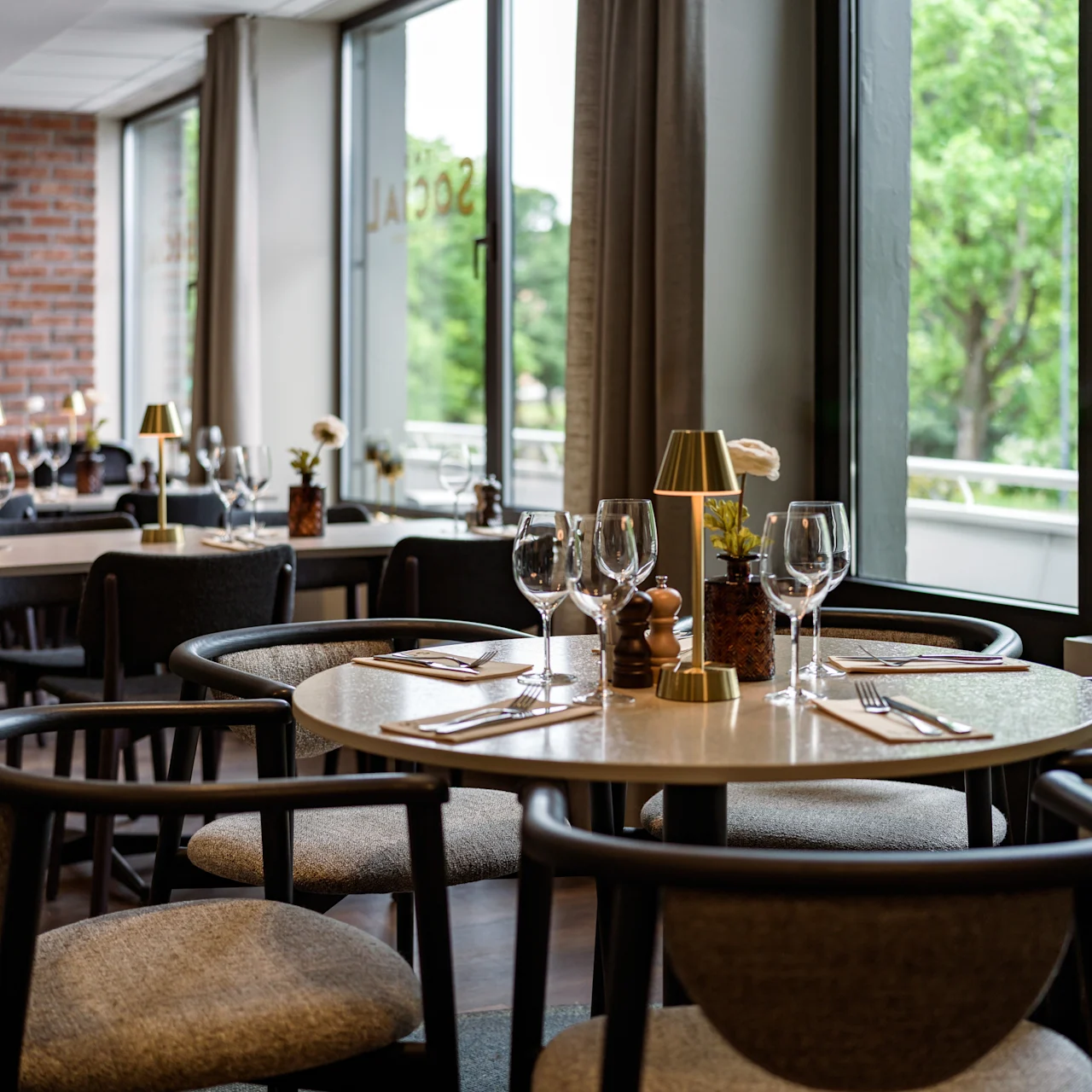 Modern dining table set with wine glasses, cutlery, and a small lamp, in a restaurant with brick walls and large windows overlooking green trees.