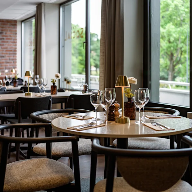 Modernt matbord med vinglas, bestick och en liten lampa i en restaurang med tegelväggar och stora fönster som vetter mot gröna träd.