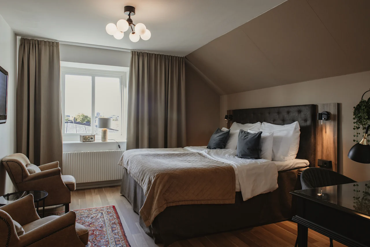 Ett mysigt hotellrum med en stor säng, två fåtöljer och ett skrivbord, badat i naturligt ljus från ett fönster.