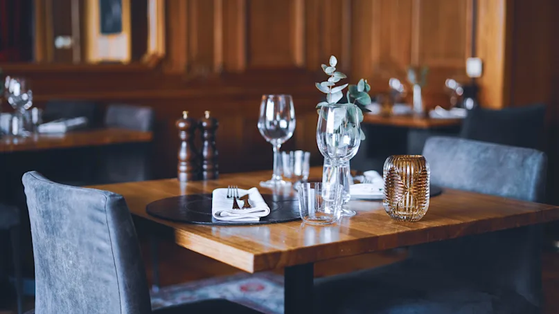 A neatly set wooden dining table with cutlery, glasses, a napkin, and a plant, conveying readiness for a meal, against a backdrop of a cozy wooden interior.