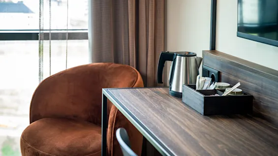 En vannkoker står på et trebord ved siden av en liten brett med kopper, nær en brun polstret stol. Innstillingen er et hotellrom med utsikt.