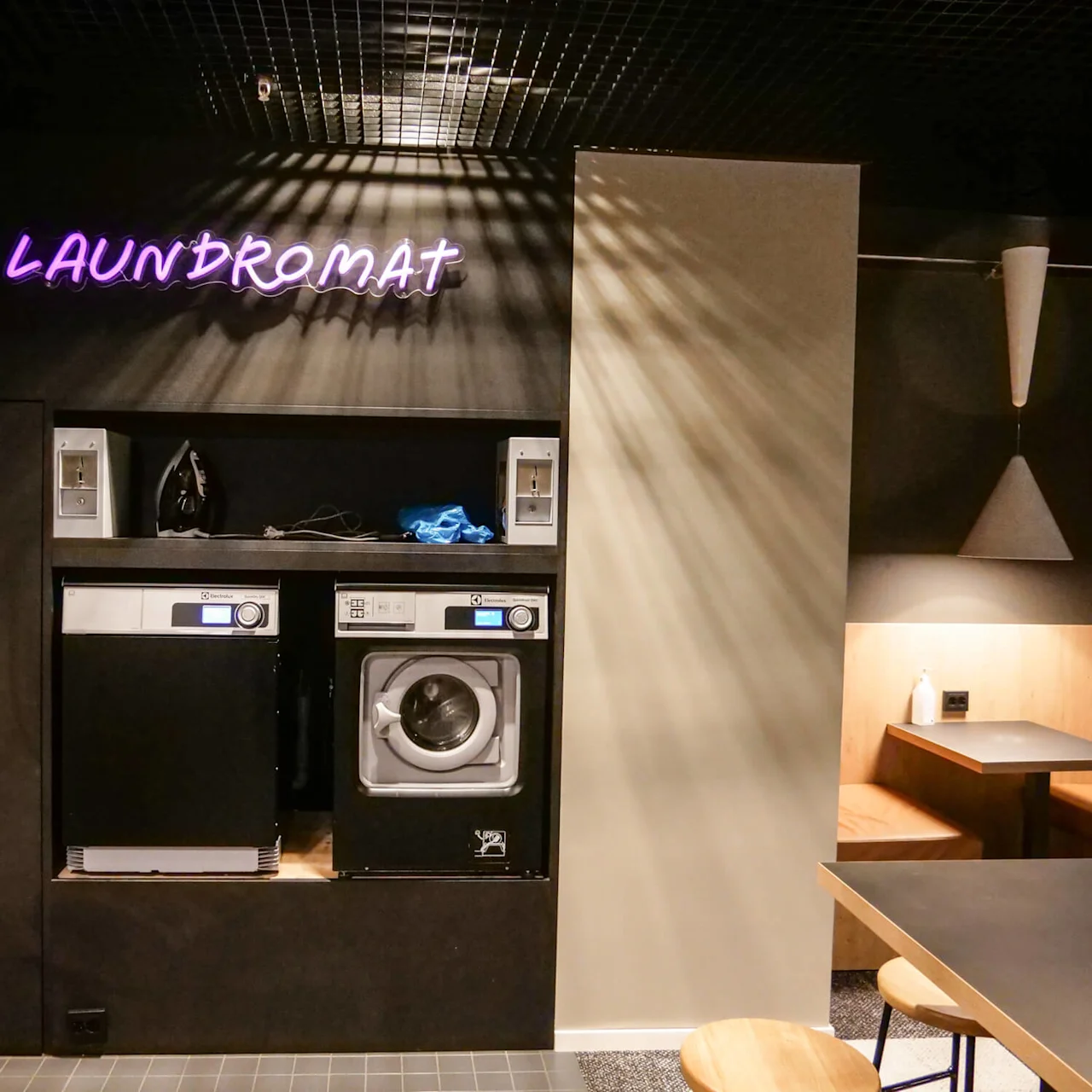 Washing machines with control panels in a modern laundromat, under neon sign reading "LAUNDROMAT." Nearby iron with colored shirt, and seating area with table and chairs.