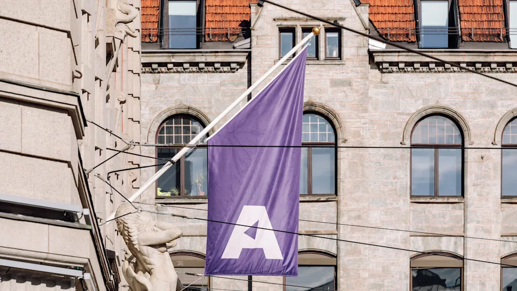 Lilla flag med et stort hvidt bogstav "A" hænger fra en bygning. Baggrund viser en stenfacade med flere buede vinduer og røde tagsten.