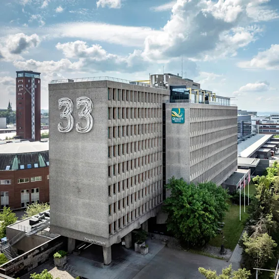 Flyfoto av en stor rektangulær bygning merket "33" og "Quality Hotel" med tilstøtende vei, trær og omkringliggende bylandskap under en delvis skyet himmel.