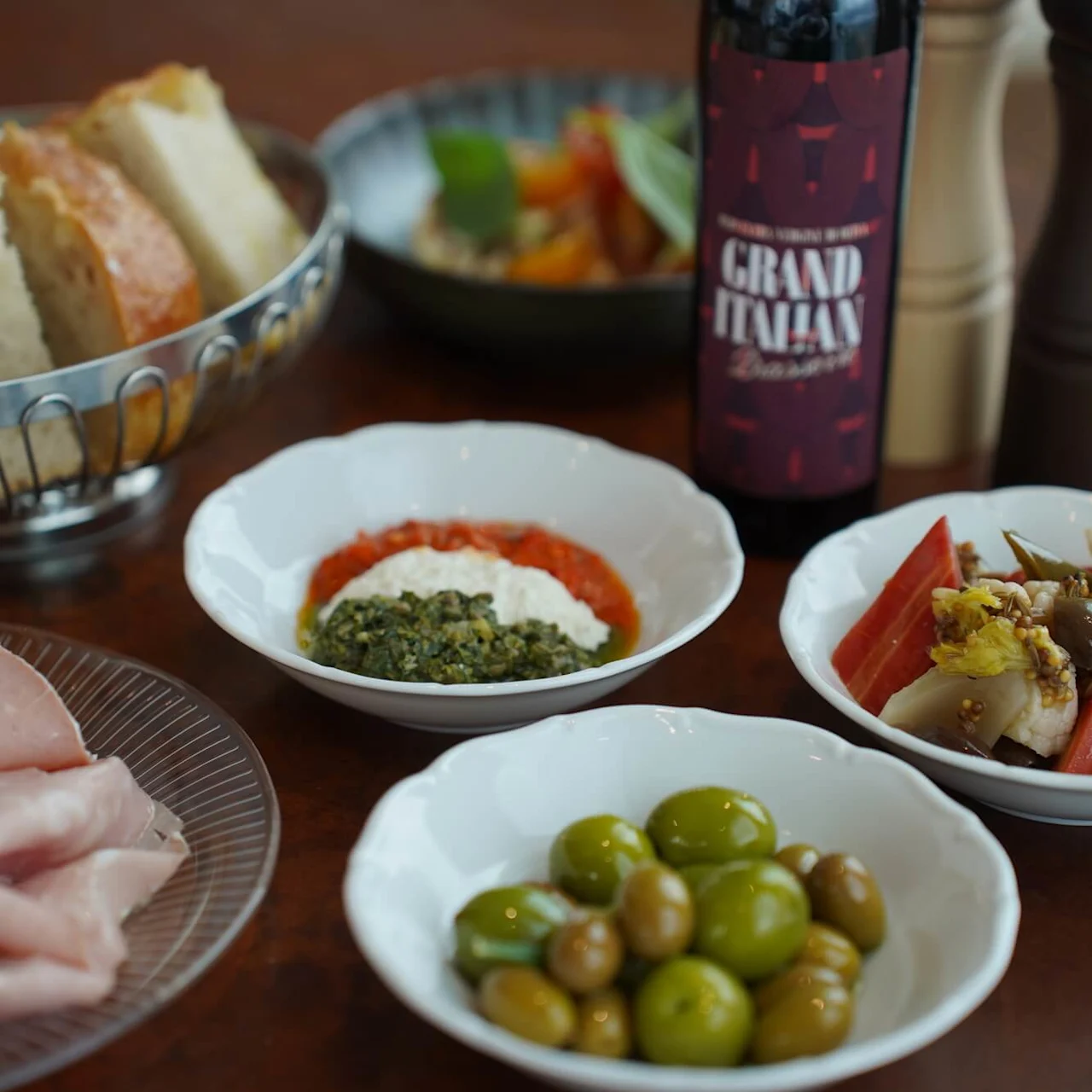 Brødskiver i en metallkurv, assorterte antipastiskåler med oliven, kjøtt og dipper på et trebord, ledsaget av en flaske merket "Grand Italian" og pepperkverner.