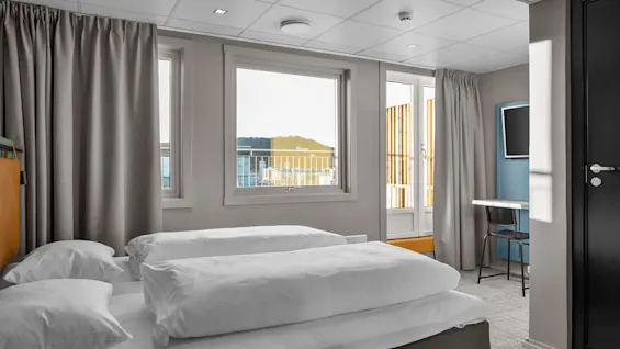 Sänky, jossa on valkoiset liinavaatteet, seisoo modernissa, valoisassa huoneessa, jossa on suuret ikkunat, harmaat verhot, pieni kirjoituspöytä ja tuoli kaupunkinäkymällä.