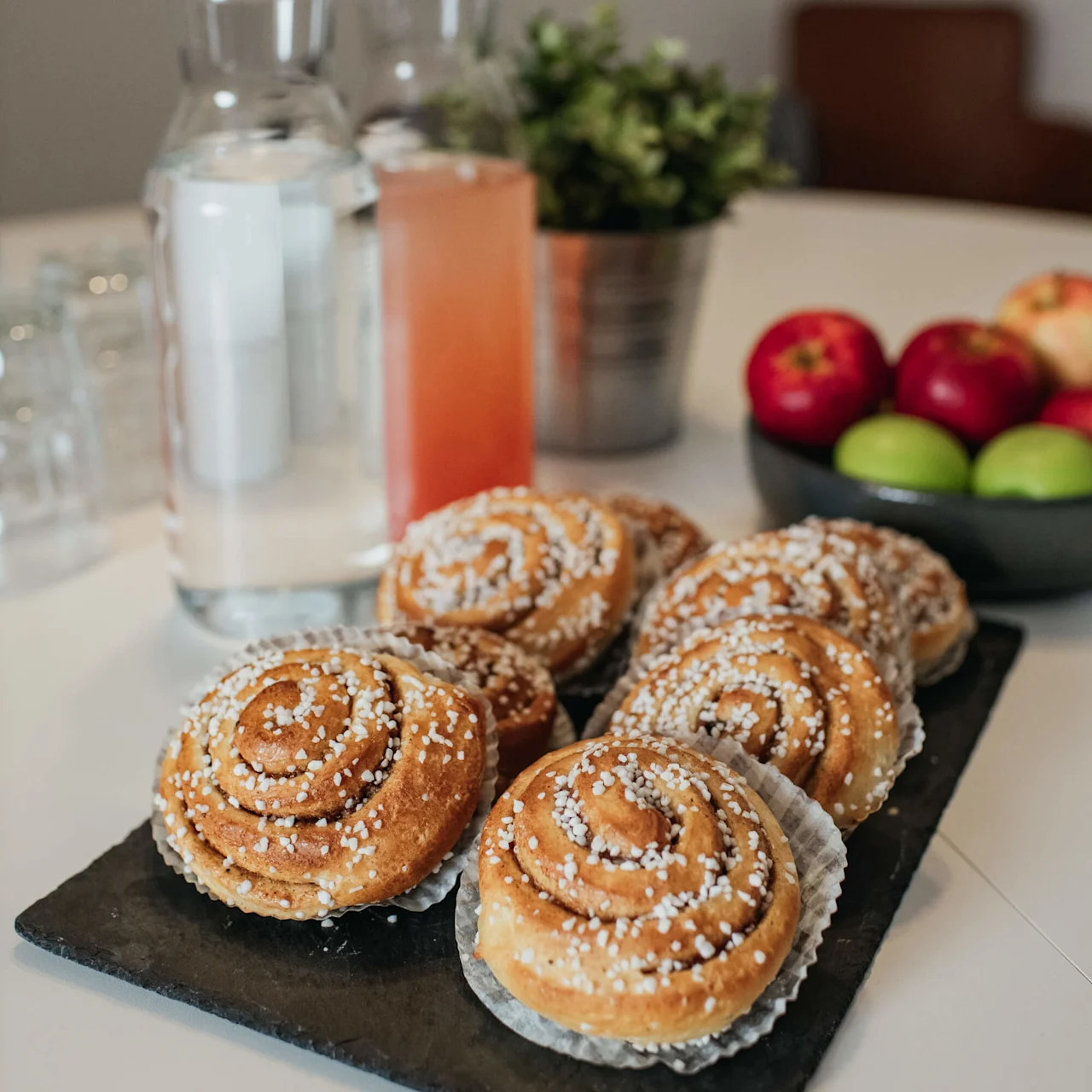 Kanelboller på en skiferplate med drikke og frukt i bakgrunnen på et bord.