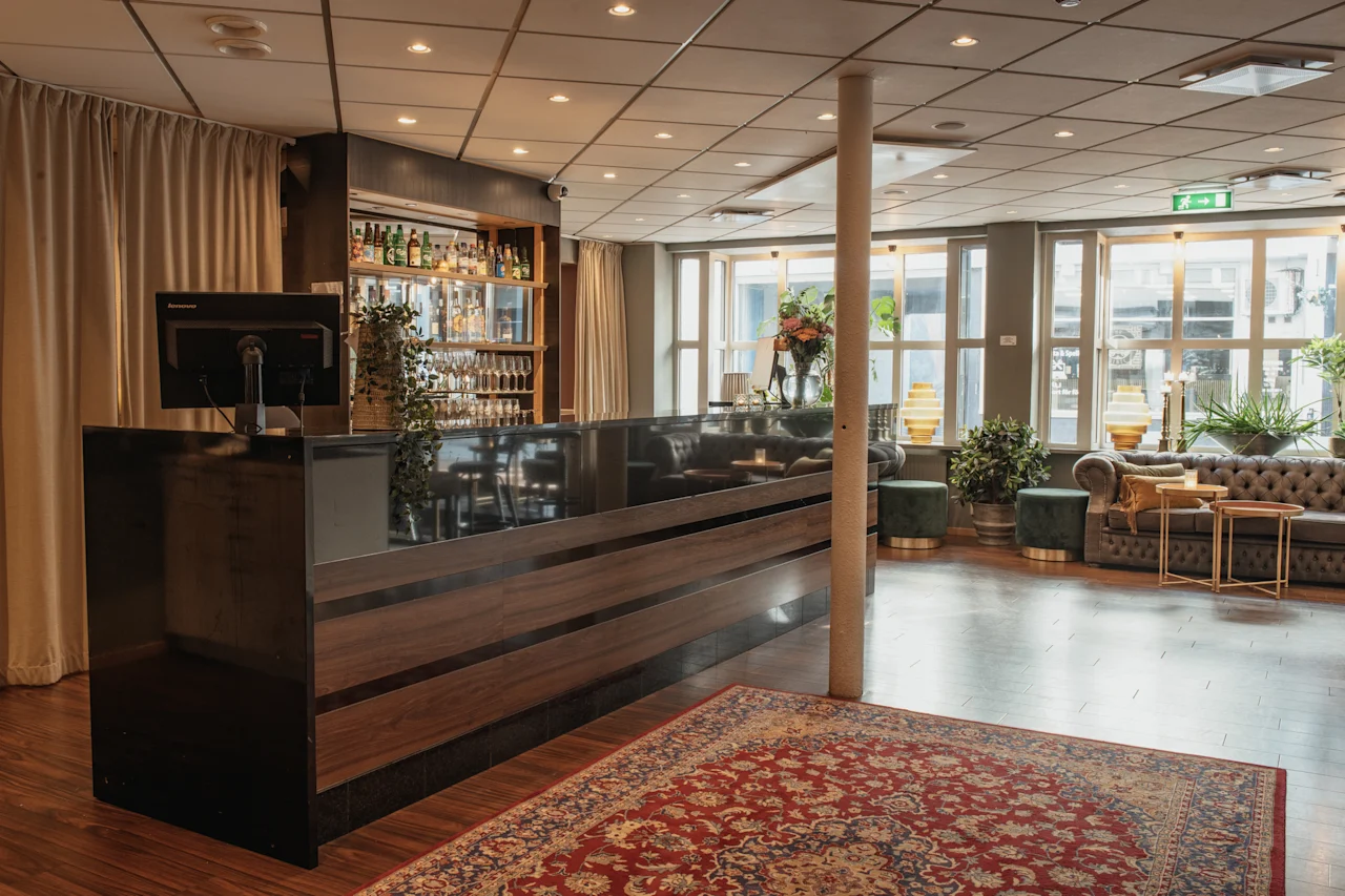 Tyylikäs hotellibaari ja lounge-alue, jossa on tumma puinen baari, mukavat istuimet, suuret ikkunat ja punainen kuvioitu matto. Poistumisnuoli.