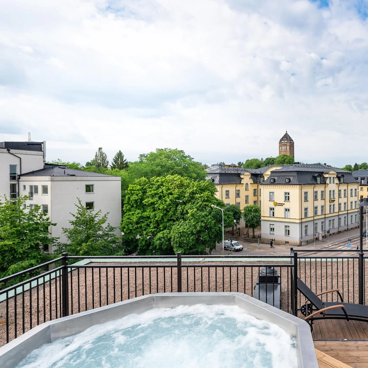 En takterrass med jacuzzi och utsikt över staden, omgiven av solstolar på ett trädäck, under en molnig himmel.