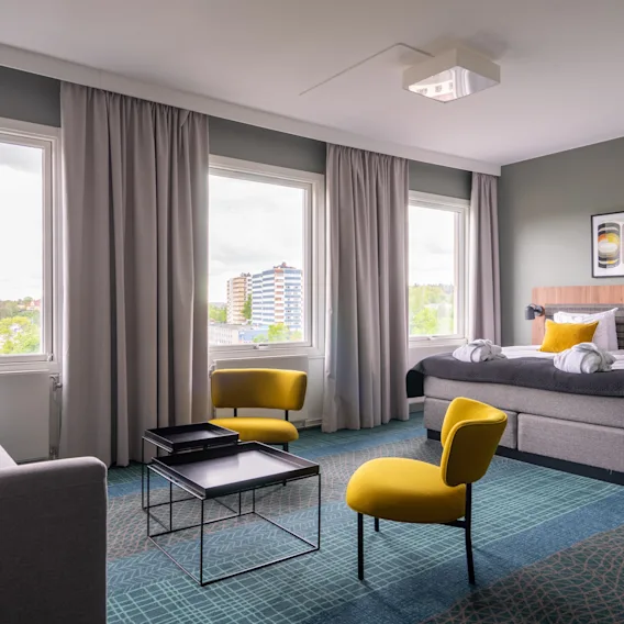 Ett hotellrum har en prydligt bäddad säng med gula kuddar, två gula stolar och en grå soffa runt ett litet bord. Stora fönster vetter mot ett stadslandskap utanför.