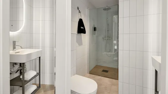 Ett modernt badrum med vita kaklade väggar, handfat till vänster, toalett i mitten och en dusch med glasdörr till höger.