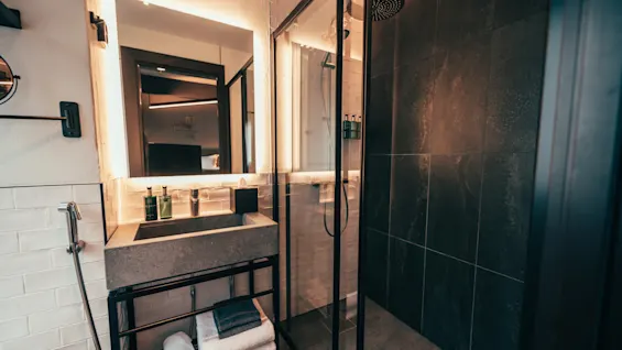 A modern bathroom featuring a sleek sink with toiletries, a brightly lit mirror, and a spacious shower with dark tiling.