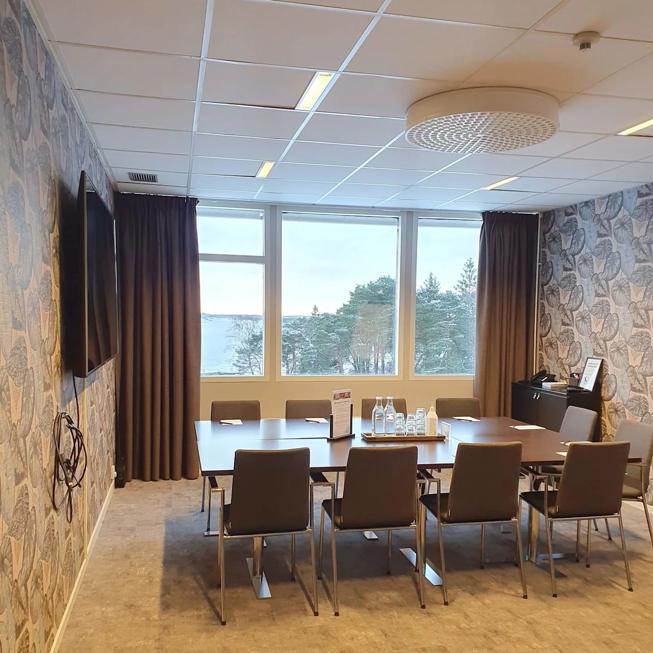 Meeting room with a wooden table and eight chairs arranged, water bottles and papers on the table. A large window overlooks trees and a lake. Walls have patterned wallpaper.