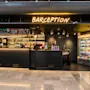Modern hotel reception and bar with neon signs reading Barception and a well-stocked deli next to it.