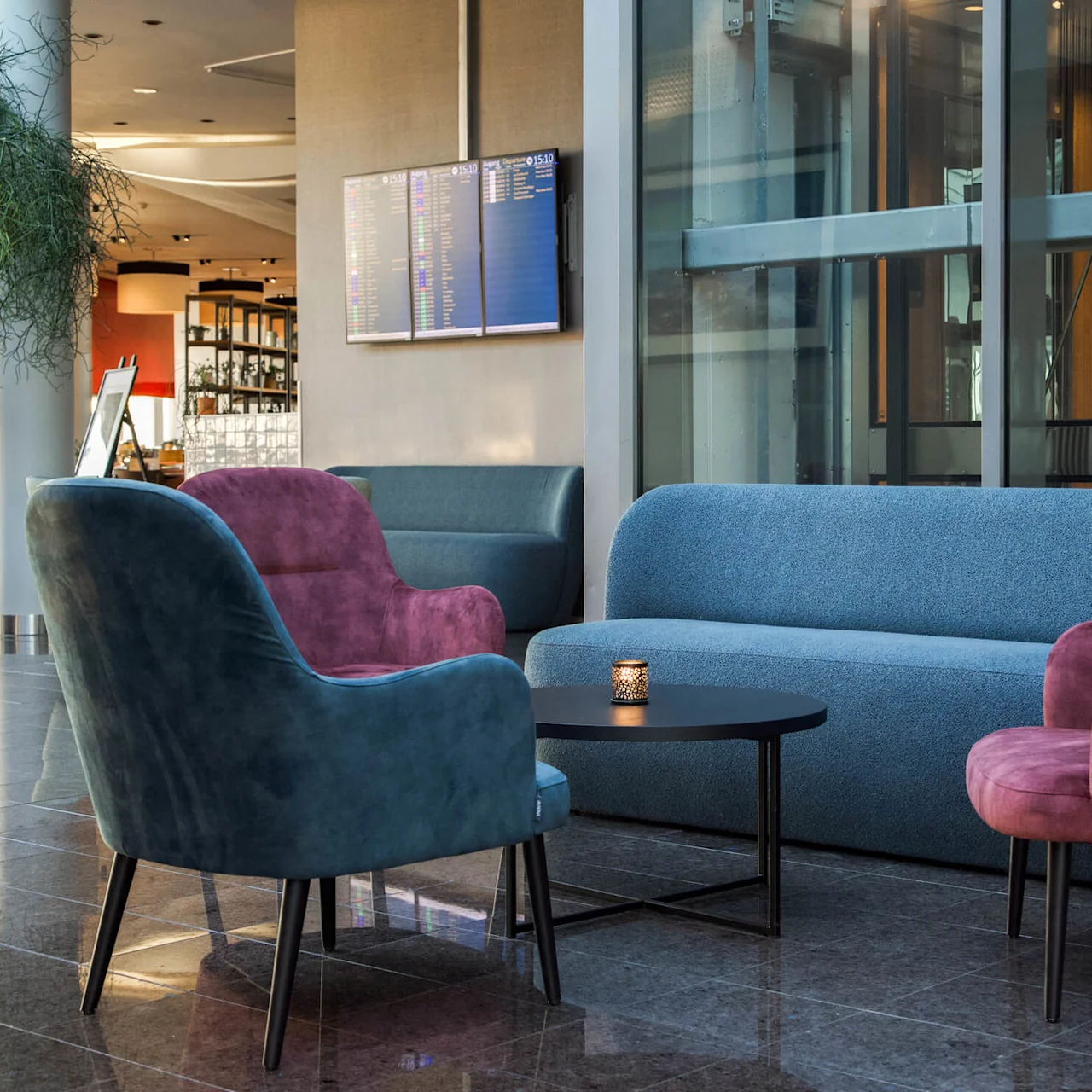 A blue sofa and pink chairs surround a black table in a lounge with flight information screens on the wall, adjacent to a modern elevator with large windows.