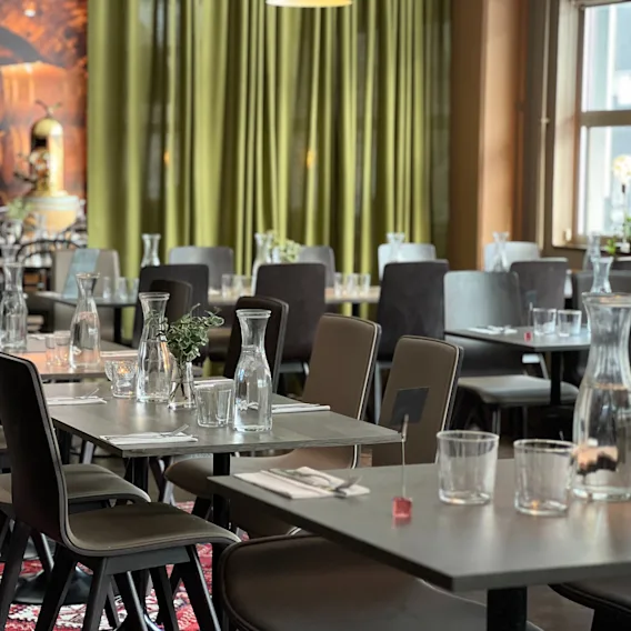 Borden är dukade med glasflaskor och koppar i en restaurang, omgivna av stolar, gröna gardiner och breda fönster, vilket skapar en rymlig och inbjudande atmosfär.