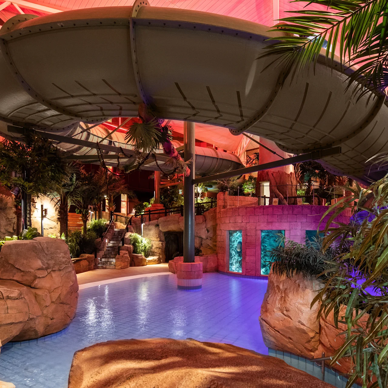 Waterslide curves over a tropical-themed indoor pool, surrounded by rocks and palm trees, creating a vibrant leisure area with colorful lighting under a high ceiling.