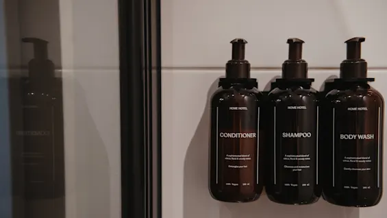 Kolme tummaa pulloa, joissa on merkinnät Hoitoaine, Shampoo ja Vartalonpesuaine, on kiinnitetty valkoiselle kaakeloidulle seinälle suihkussa.