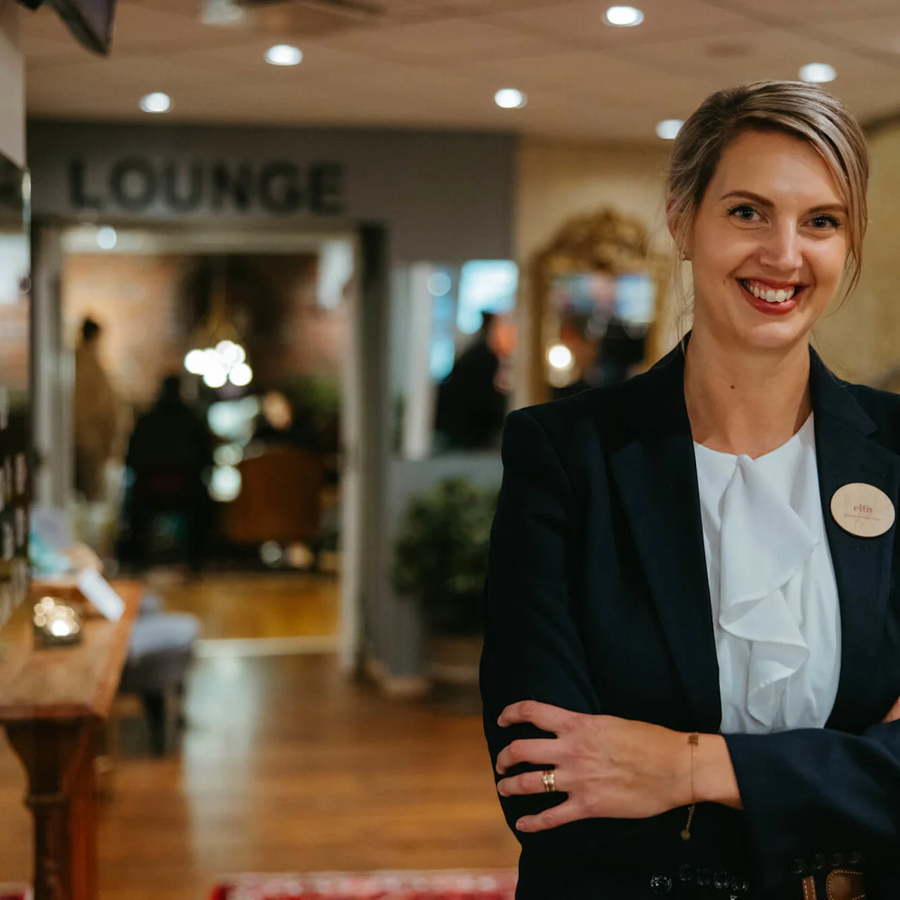 Hymyilevä nainen liikepukeutumisessa aulassa, nimilaatta tekstillä "Cath", taustalla suttuinen kyltti "LOUNGE".