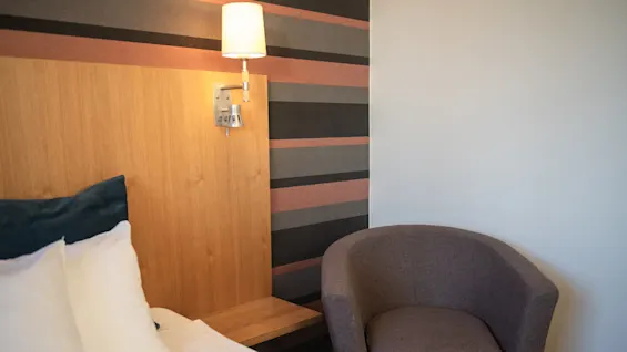 Bed with white pillows beside a brown armchair, under a lit wall lamp on wooden paneling. Striped wall design in the background.