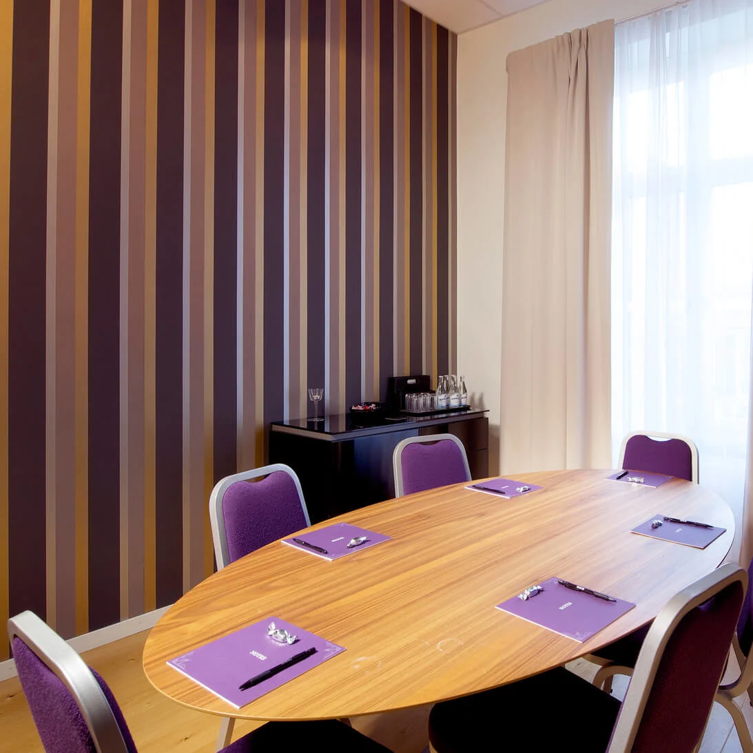 A modern meeting room with a wooden table, purple chairs, and striped wallpaper, lit by natural light streaming through a window.