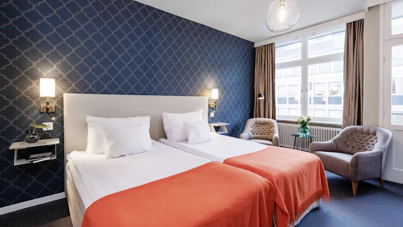 A neatly presented hotel room with a patterned blue wallpaper, a large bed with white bedding and an orange throw, flanked by side tables and lamps, and comfortable seating near a window with curtains.