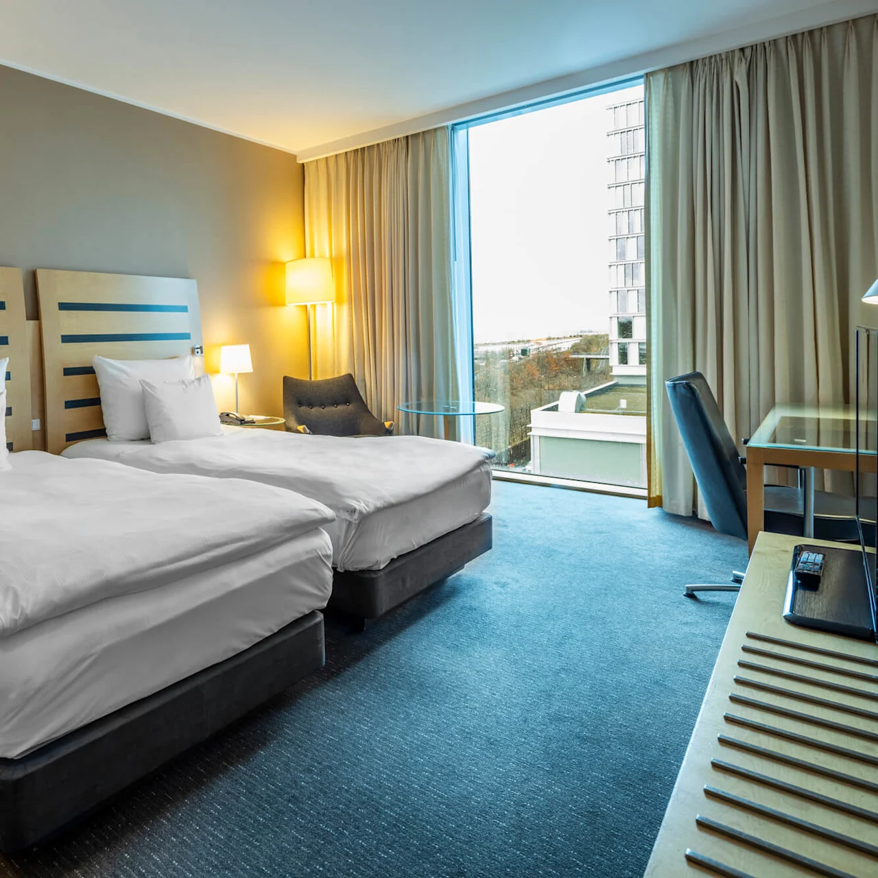 Två prydligt bäddade enkelsängar står sida vid sida, omgivna av lampor, i ett modernt hotellrum med stort fönster, skrivbord och stol.