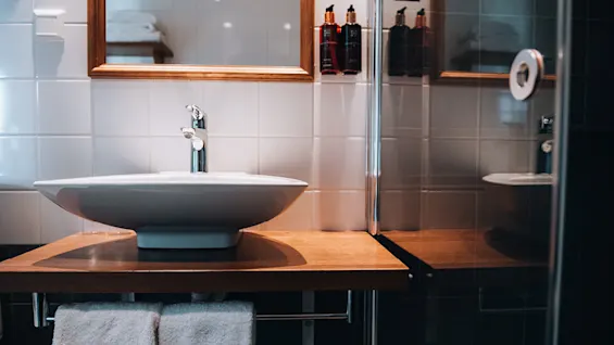 En moderne håndvask med en stilfuld hane står på en træbordplade. Flisevægge og et spejl med reflekterede toiletartikler fuldender det sofistikerede miljø.