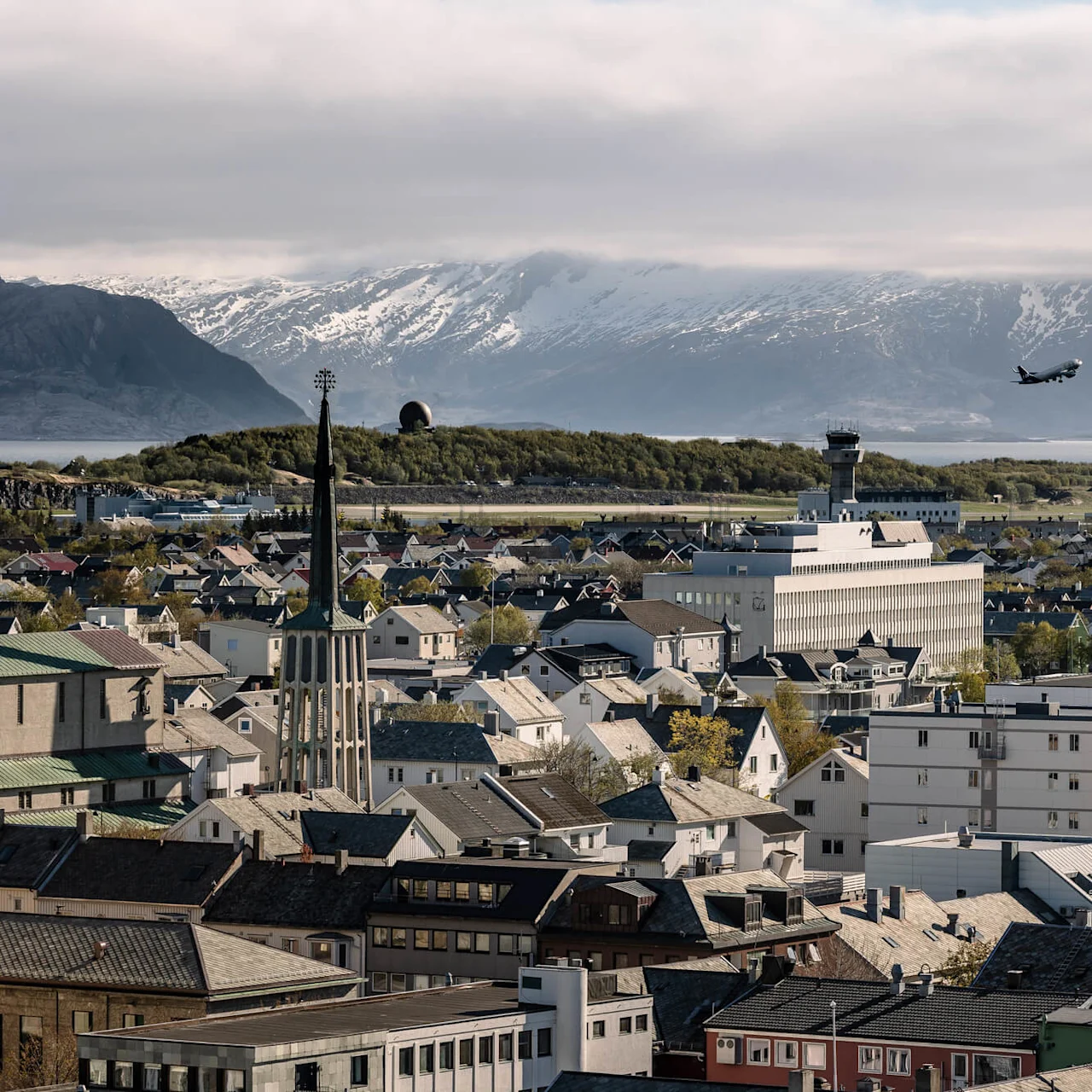A plane ascends above a cityscape, featuring a mix of modern and traditional architecture, with snow-capped mountains and a cloudy sky in the background.