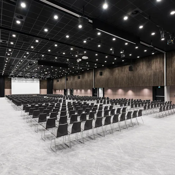 Rader av svarte stoler er arrangert i et stort auditorium, vendt mot en hvit prosjektor. Lyse taklys og mørke trevegger skaper et moderne og romslig miljø.