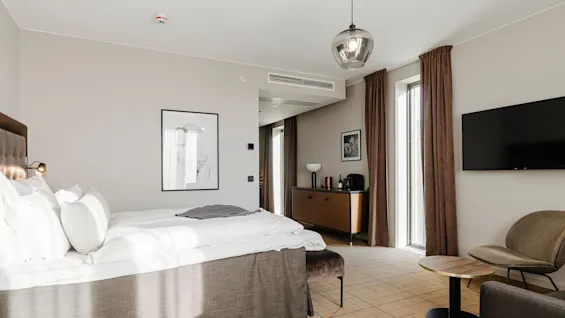 Et moderne hotelværelse har en stor seng med hvidt sengetøj. En sengelampe er fastgjort til væggen. Et skab har en flaske og kaffemaskine nær et vindue med brune gardiner.