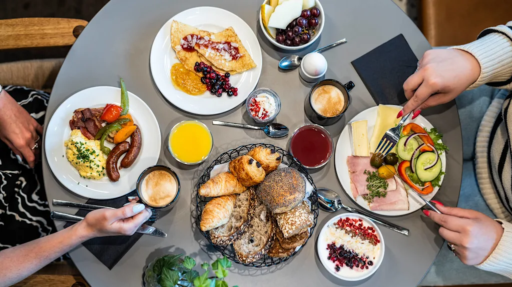 An overhead view of a beautifully set breakfast table featuring fresh bread, croissants, yogurt with berries, scrambled eggs, sausages, cheese, fresh vegetables, juice, coffee, and fruits.
