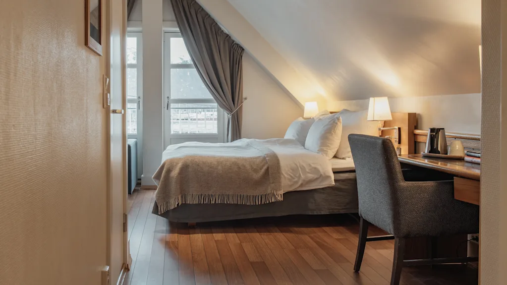 A cozy hotel room with a comfortable bed, a desk, and a chair, featuring a sloped ceiling and a window with curtains.