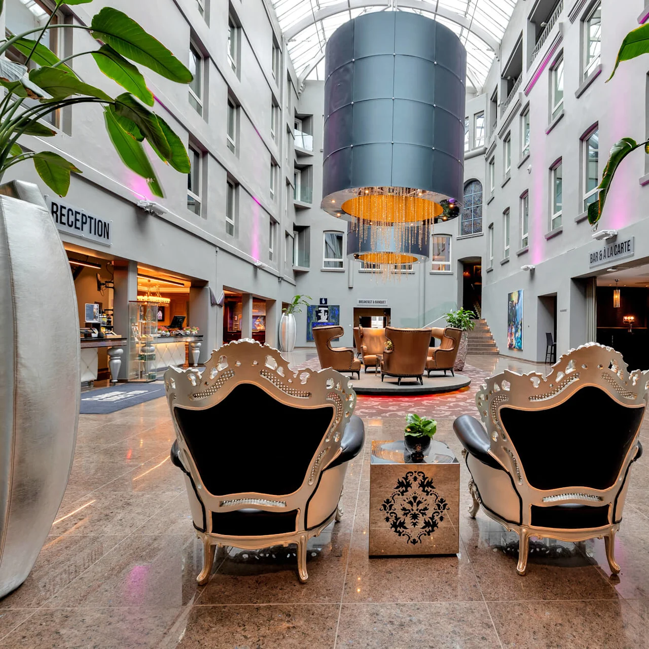 To udsmykkede stole vender mod et loungeområde under en stor lysekrone i et rummeligt atrium. Skilte siger "RECEPTION" og “BAR & A LA CARTE.” Høje planter indrammer scenen.