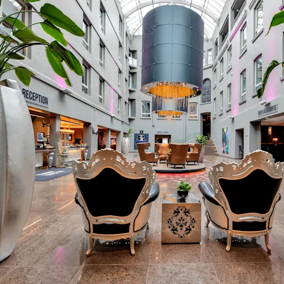 Two ornate chairs face a lounge area under a grand chandelier in a spacious atrium. Signs read ”RECEPTION” and “BAR & A LA CARTE.” Tall plants frame the scene.
