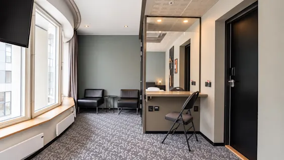 Bright hotel room featuring a desk, mirror, chairs, and window.