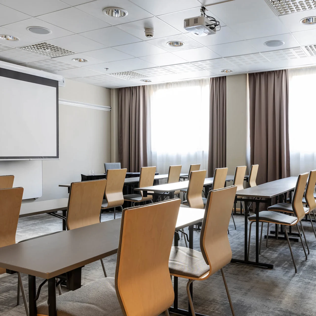 Classroom with empty wooden chairs arranged around long tables, facing a large projection screen. Room is well-lit with natural light from windows covered by brown curtains.