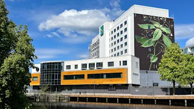En fler-våningsbyggnad visar en stor botanisk väggmålning på fasaden. Den står bredvid ett lugnt vattendrag, omgivet av frodiga gröna träd, under en klarblå himmel. Text: "SCANDIC".